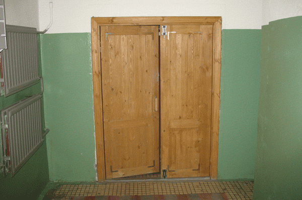 Деревянная глухая двустворчатая дверь установлена в парадной современной многоэтажки. Одно полотно закрывается на шпигалеты, второе рабочее.